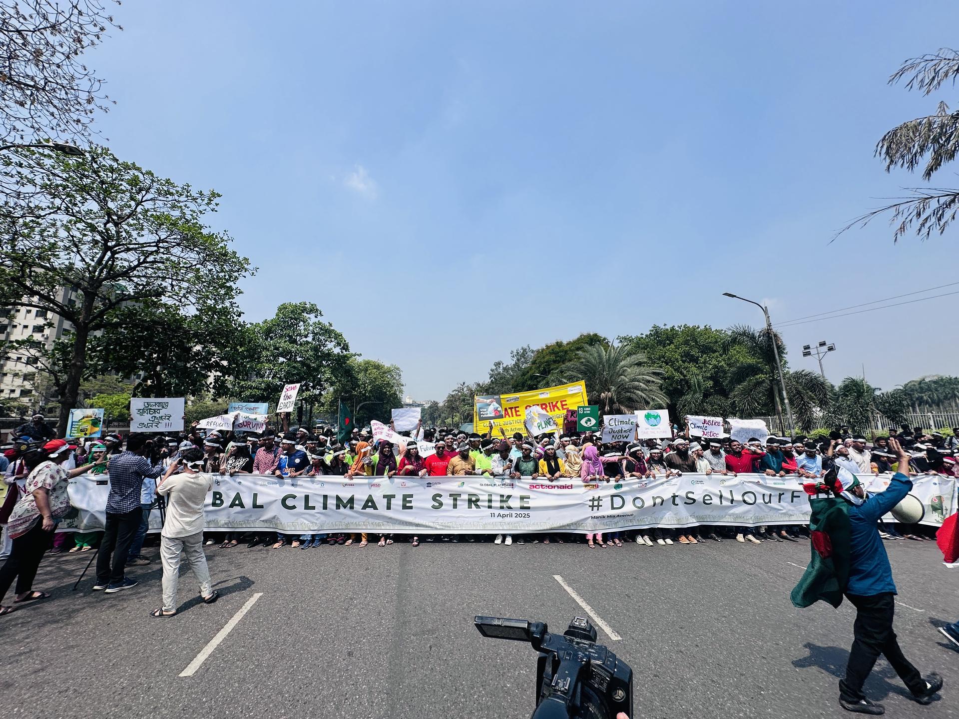 Large crowd of protesters marching on a sunny street with signs and a banner; camera in foreground.