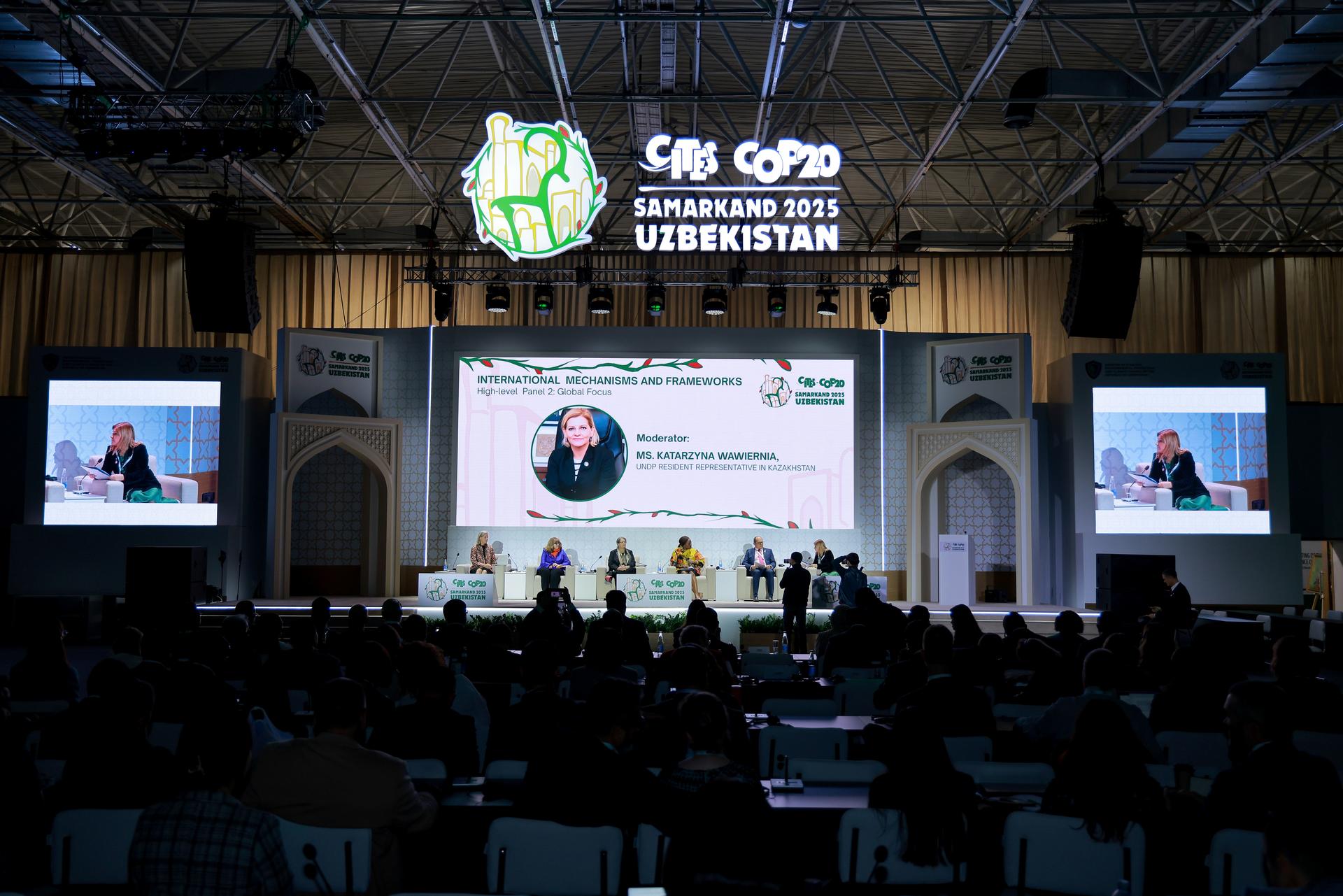Conference hall hosting a panel discussion; large logo on screen and audience.