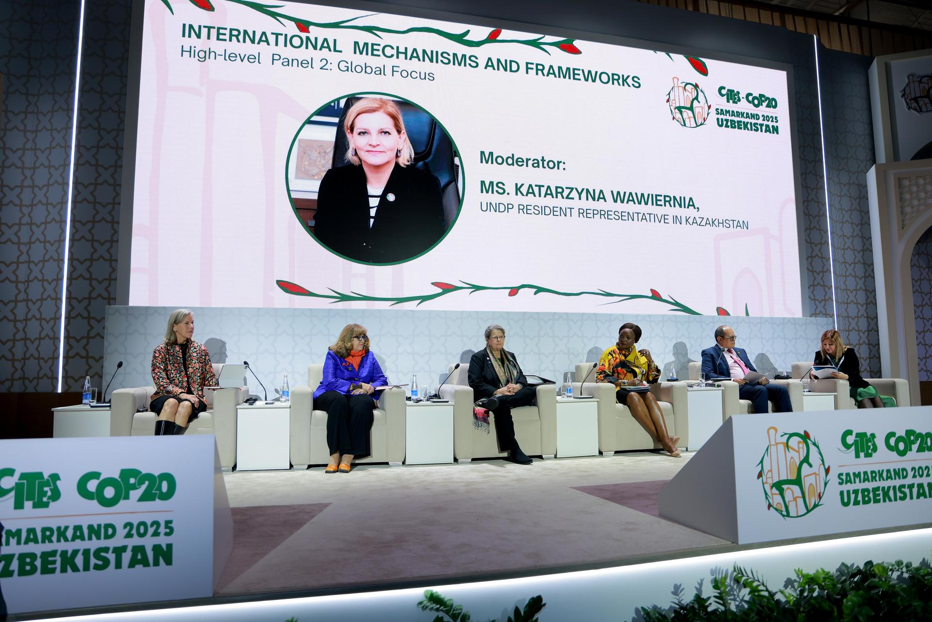 Conference stage with five panelists and a large display screen behind them.