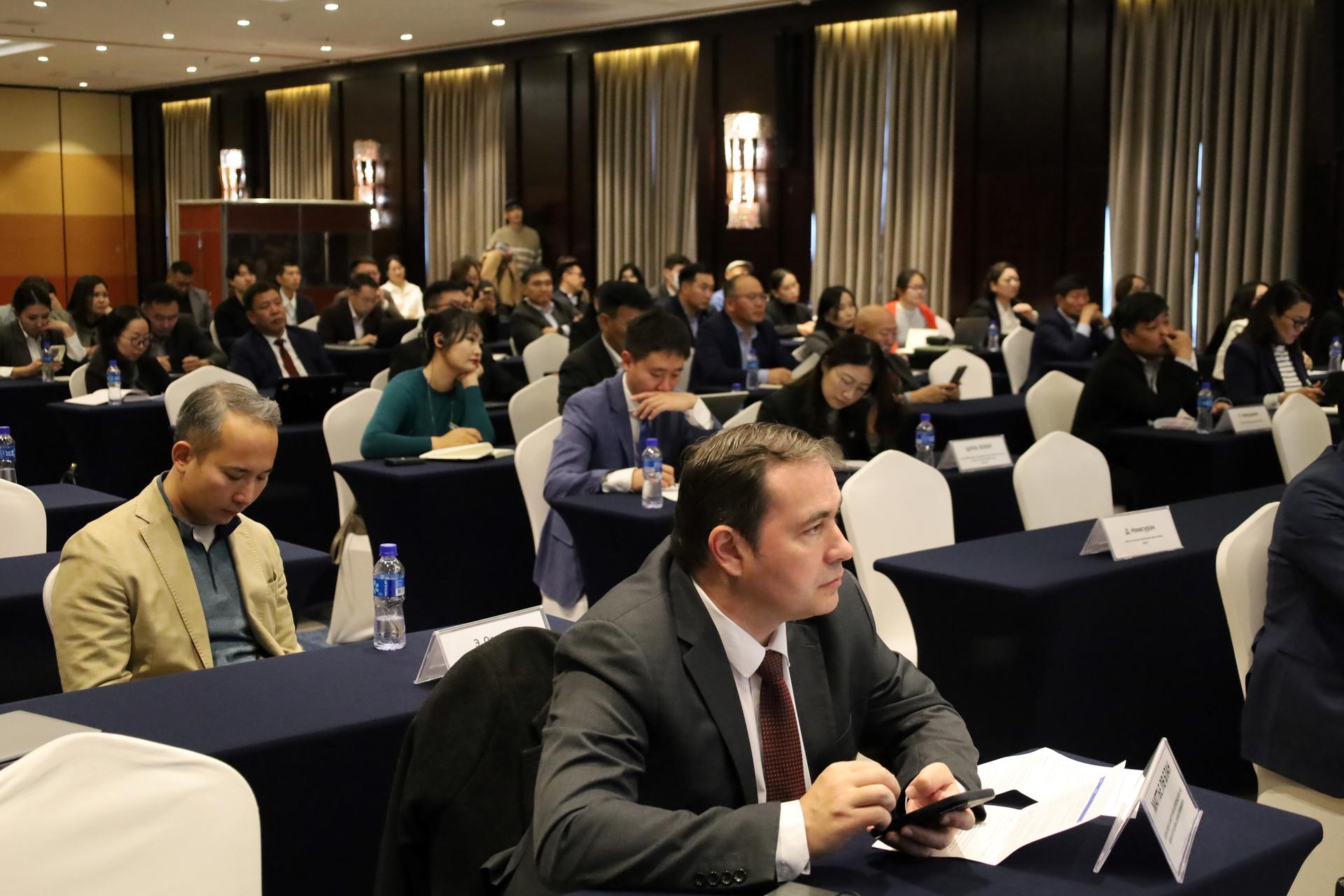 Photo: conference room filled with businesspeople at tables; foreground man in suit taking notes.