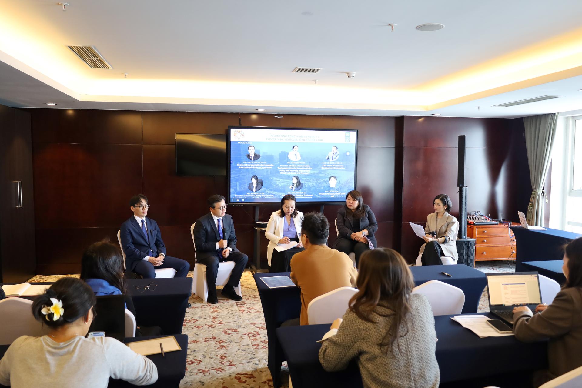 Panel discussion in a conference room with attendees around tables and a presentation screen.
