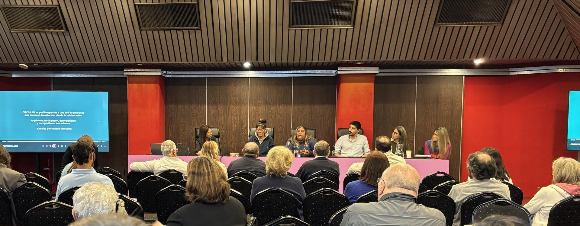 Panelists on stage in a conference, addressing an audience in a red-walled lecture hall.