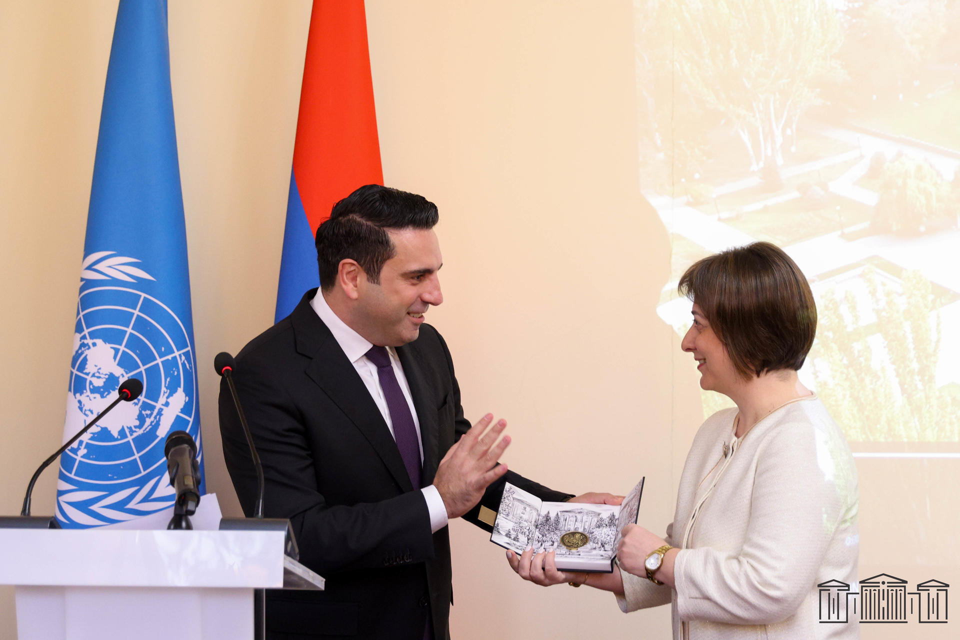 Man in suit presents award to woman in white blazer beside UN flag and podium.