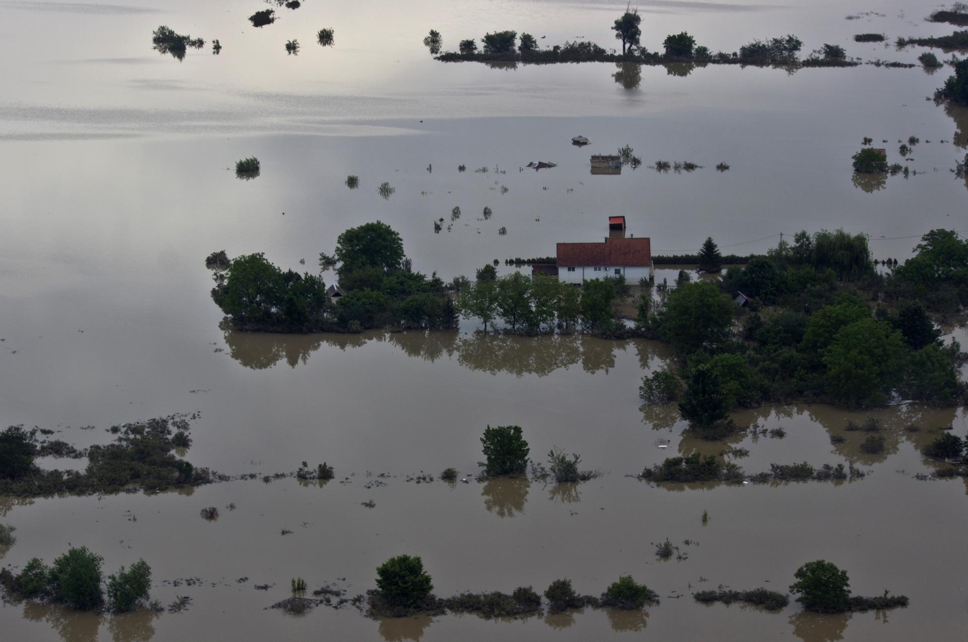 Flooded rural landscape with partially submerged trees and a red-roofed building under gray sky.