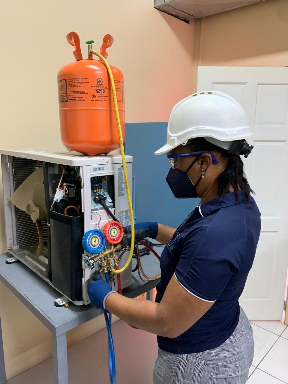 Technician in hard hat and mask servicing HVAC unit with gauges and orange refrigerant tanks.