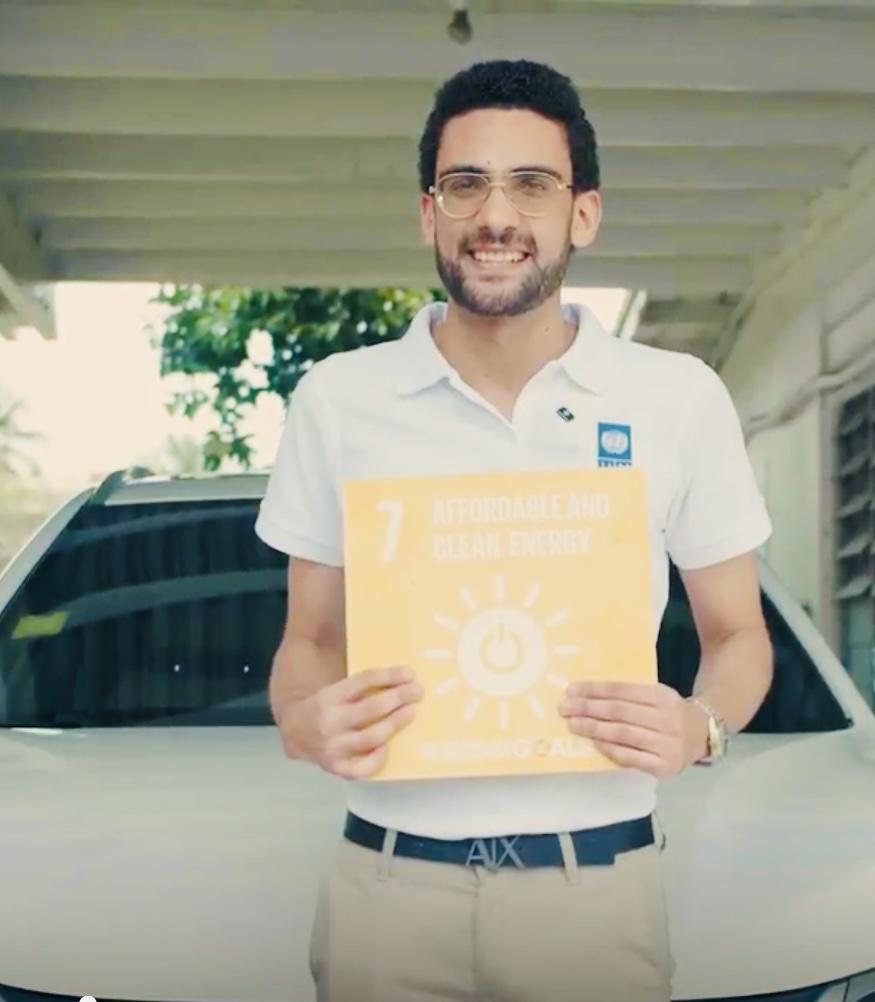 Photograph of a man in a white polo holding an orange booklet with a sun icon, in front of a car.