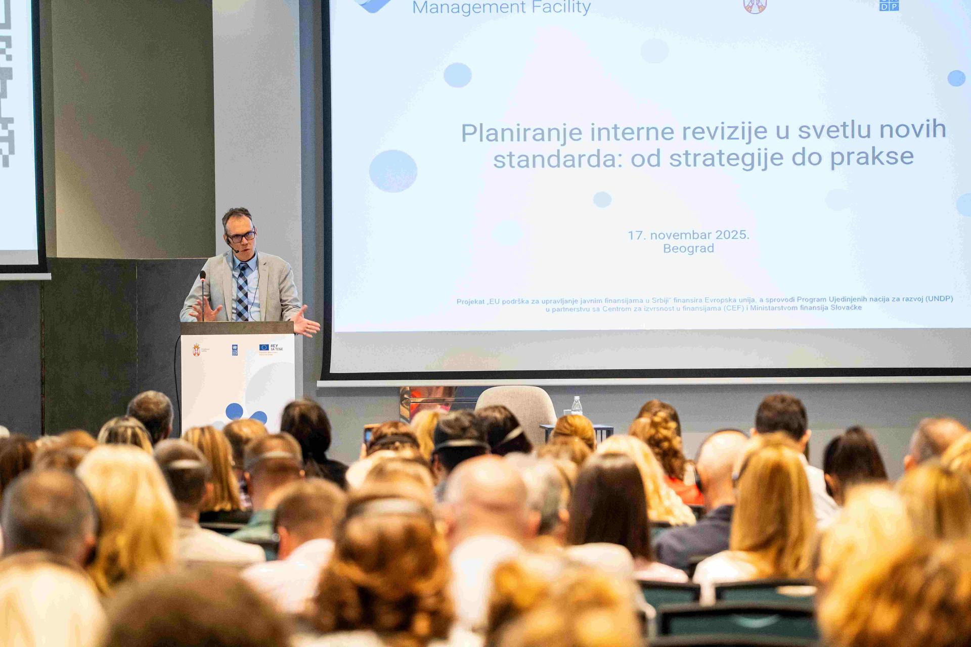 Photo of a speaker at a podium presenting to an audience in a conference hall with a slide.