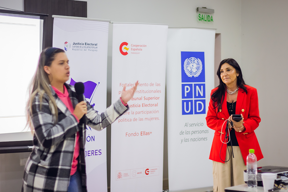 Two women on a conference stage; one gestures, the other speaks, banners with logos behind.
