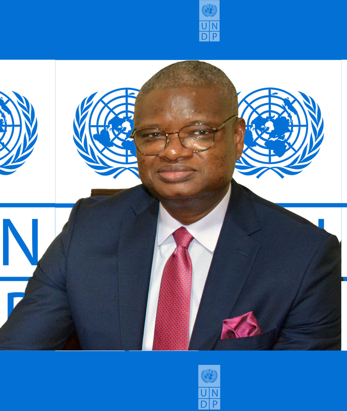 Professional in navy suit with pink tie and pocket square, seated before United Nations logos.