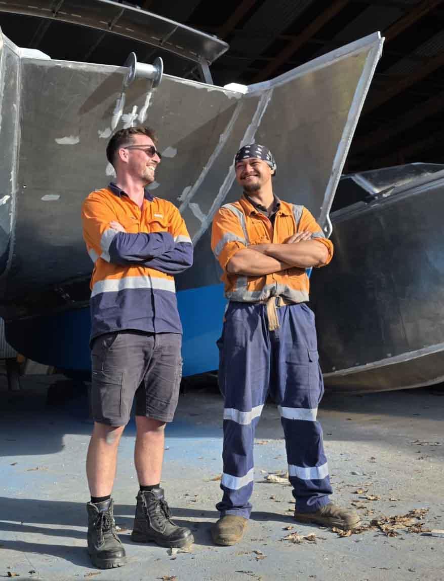 Two workers in orange shirts stand with arms crossed in front of a large ship hull at a dry dock.