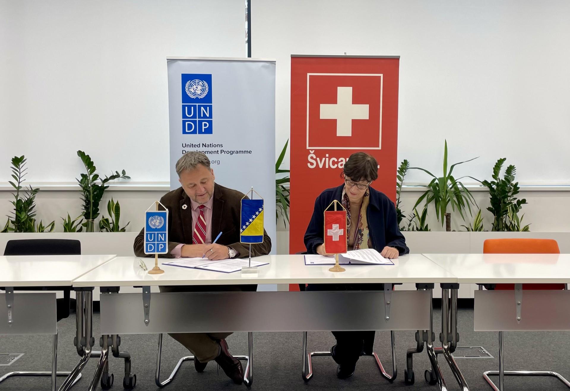 Two speakers at a conference table with blue and red banners behind them, featuring a Swiss flag.