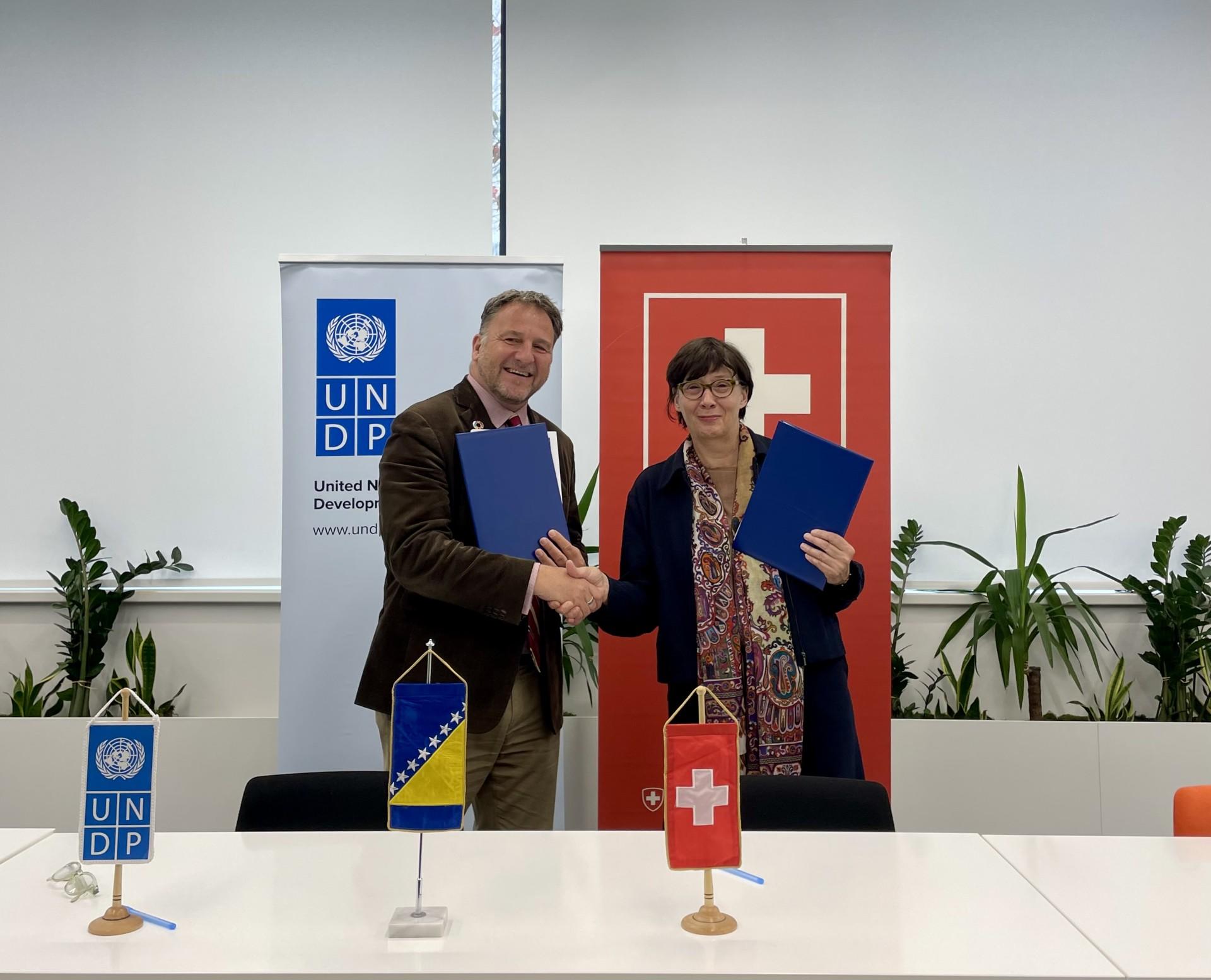 Two people sign documents and shake hands at a signing ceremony; UNDP banner and flags visible.