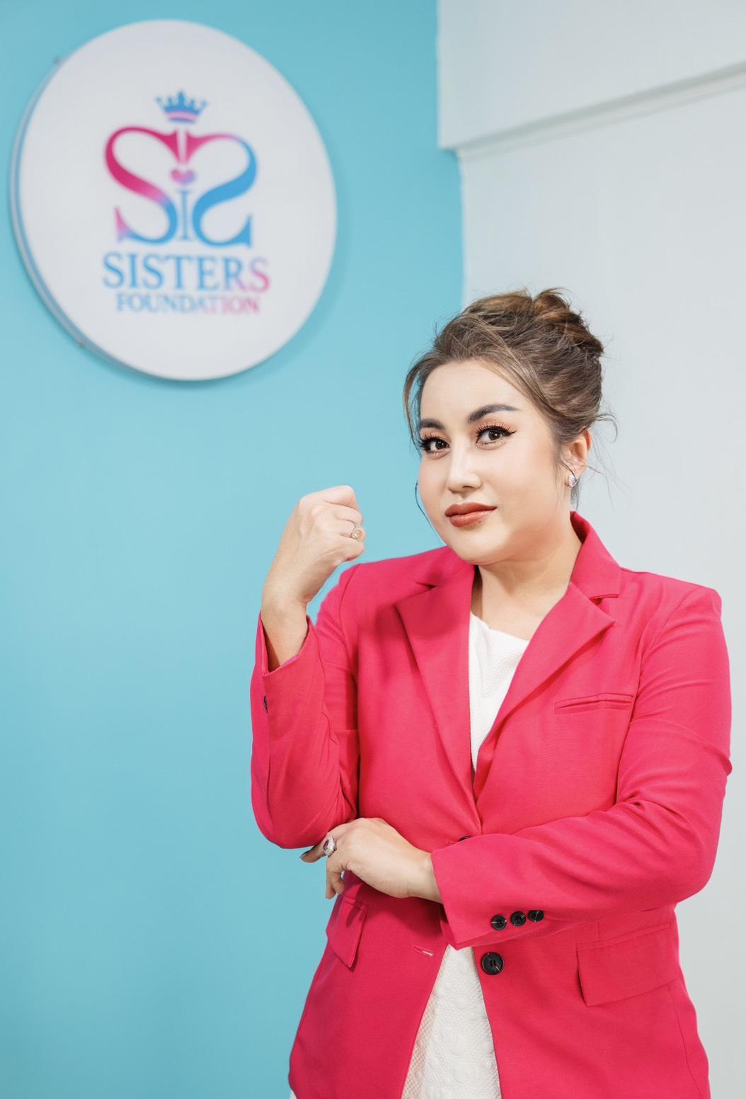 Woman in a pink blazer stands against a blue wall with a circular Sisters logo.