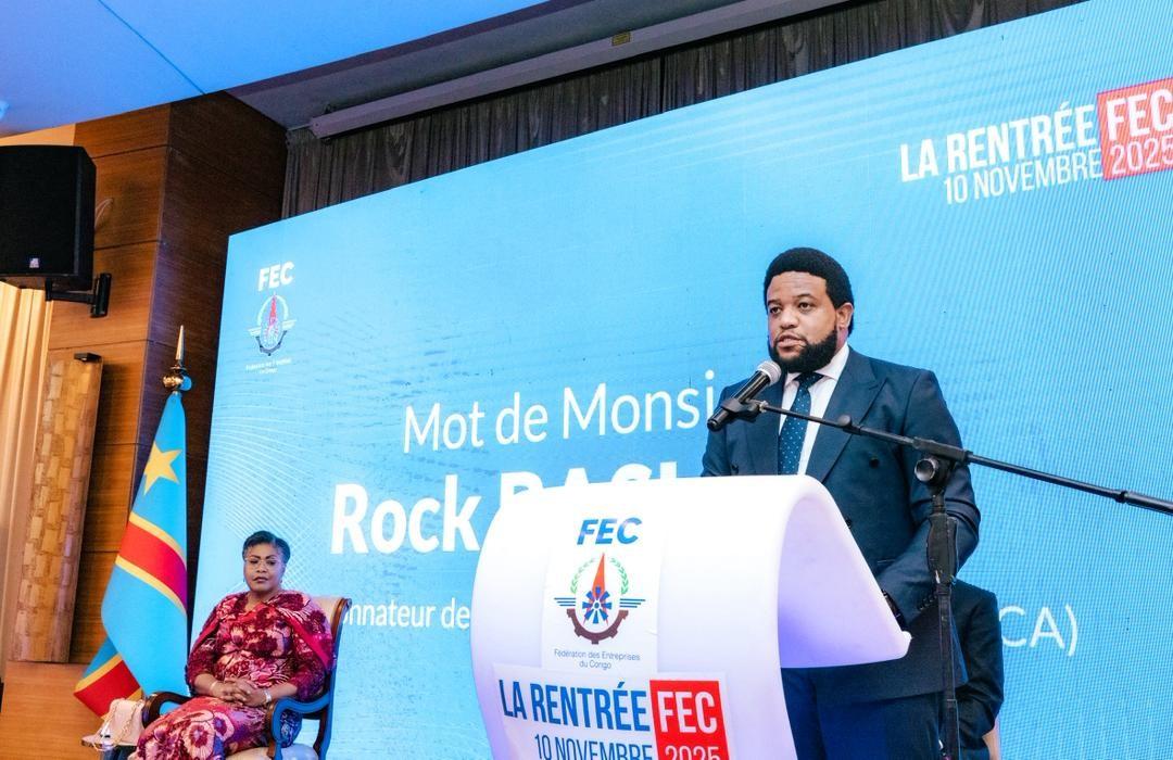 Male speaker at a podium during a conference; blue backdrop with logos, seated woman in red left.