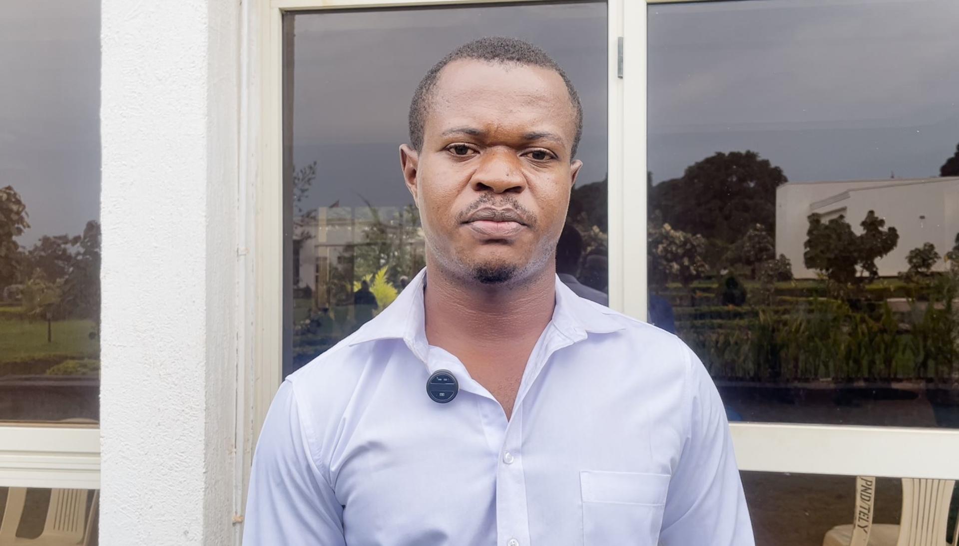 Man in a light blue button-up shirt stands outside in front of a window, with trees behind.
