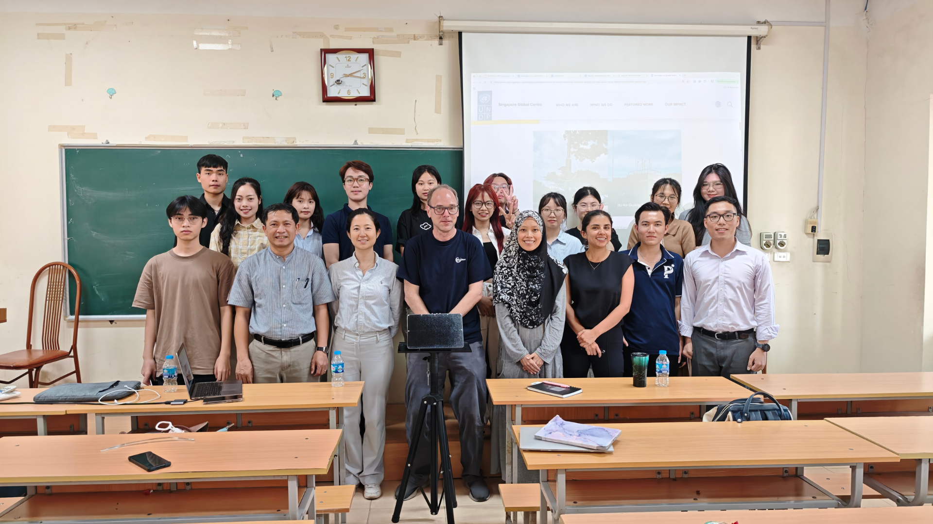 A group photo of 19 participants standing in two rows at the front of a classroom, with a sensor in the middle.