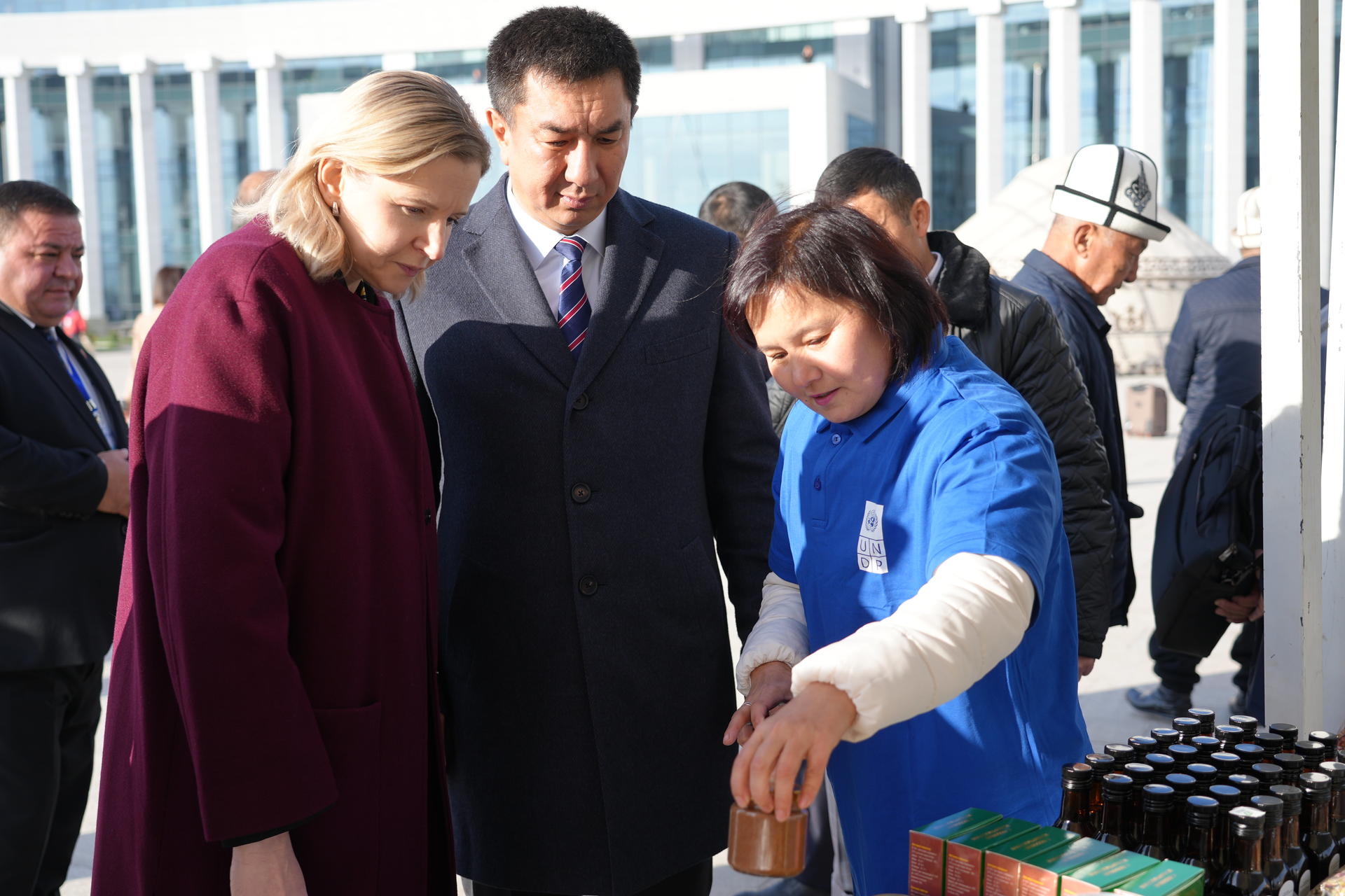 People at an outdoor market; a person in a blue jacket points at bottles on a table.