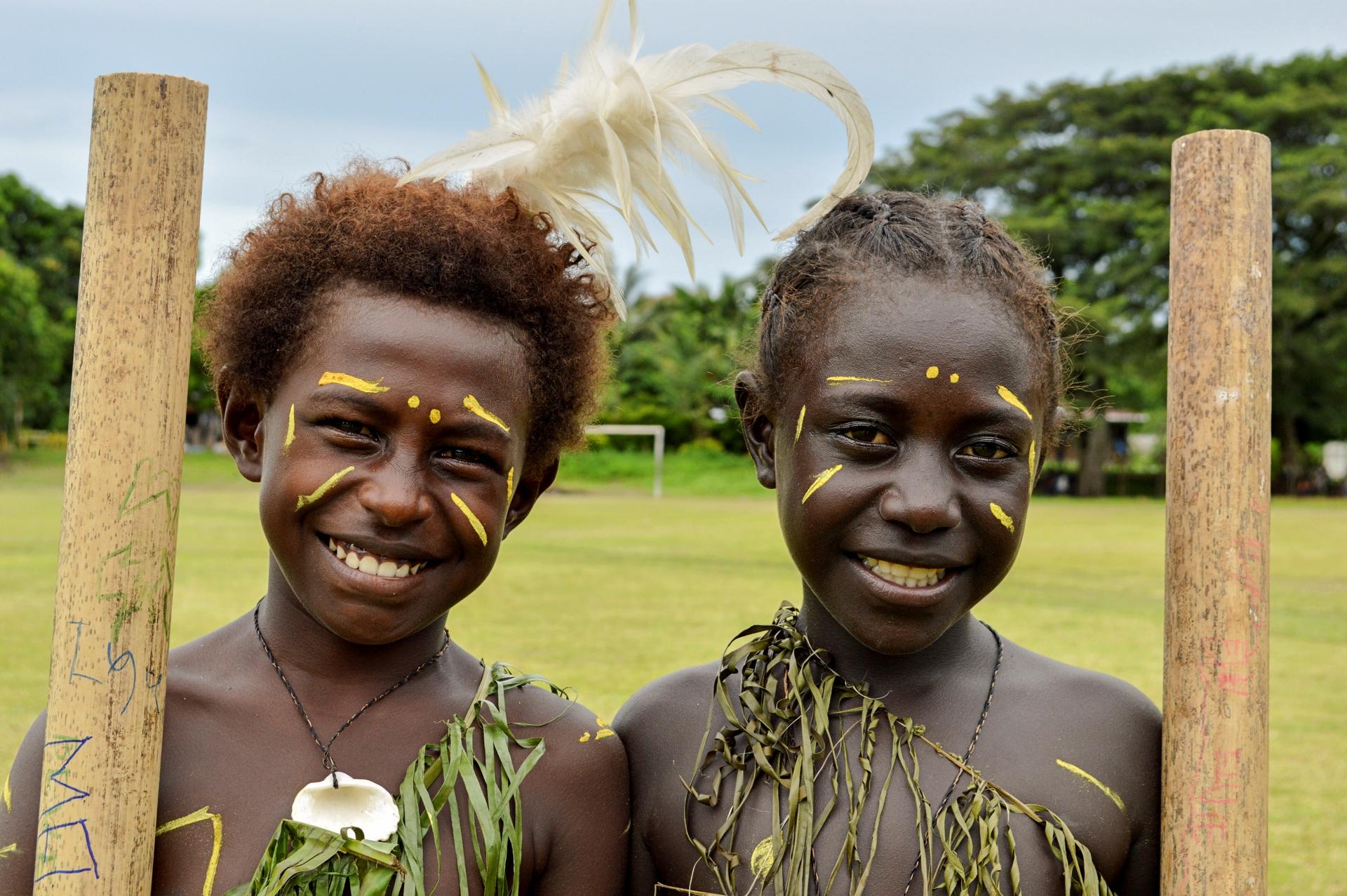 Two people in grass skirts and shell necklaces pose outdoors beside wooden posts.