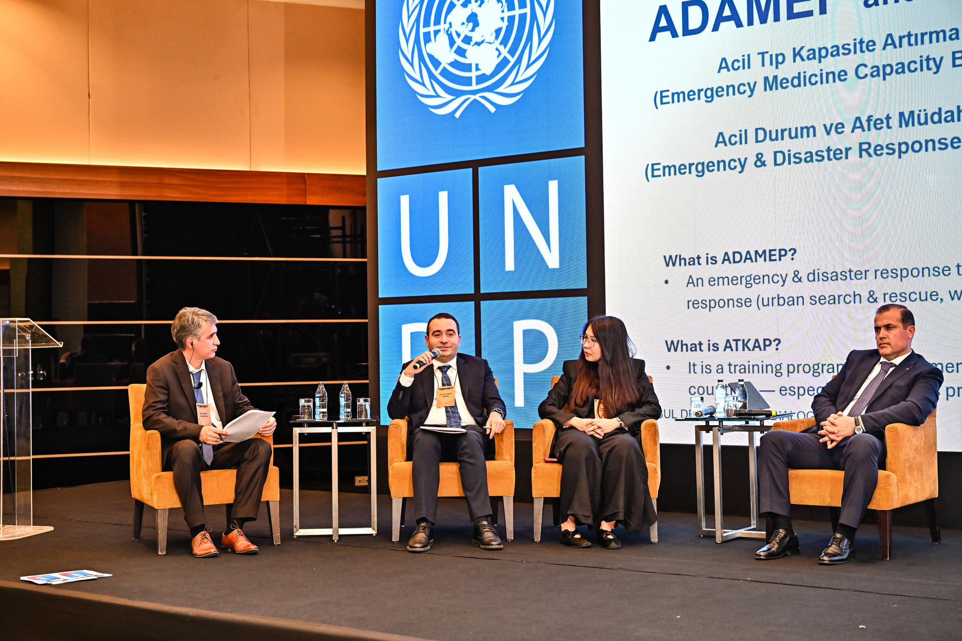 Five panelists sit in orange chairs on a stage with UN backdrop and banner.