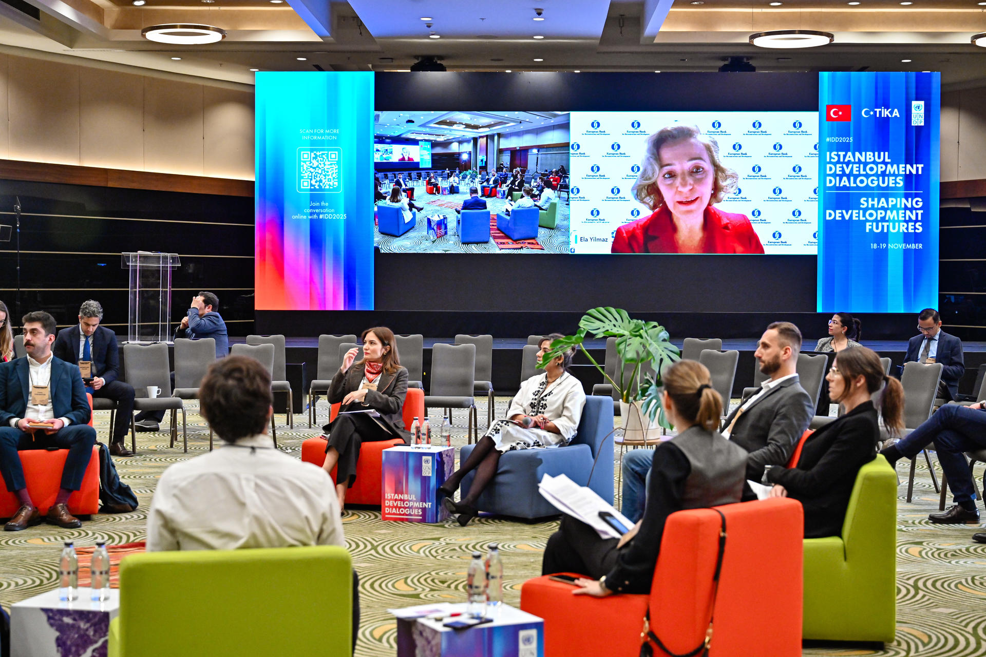 Photograph of a conference hall with colorful chairs and a large screen showing a speaker