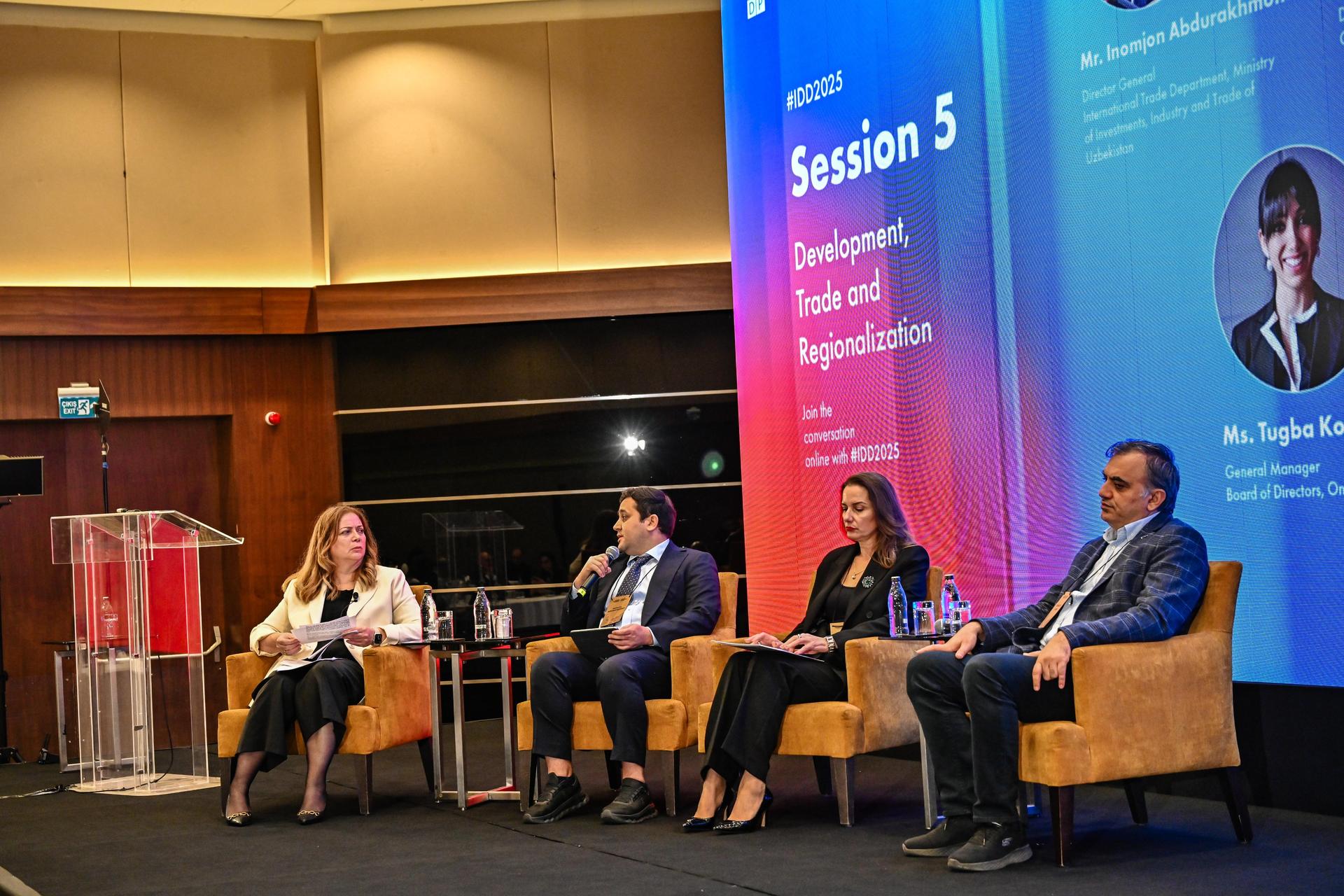 Panel discussion on stage with five speakers seated in chairs; blue backdrop reads Session 5.