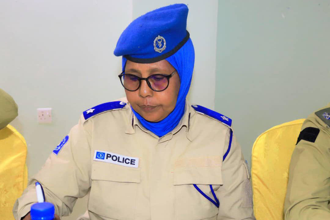 Policewoman wearing uniform with blue beret and POLICE patch, seated indoors.