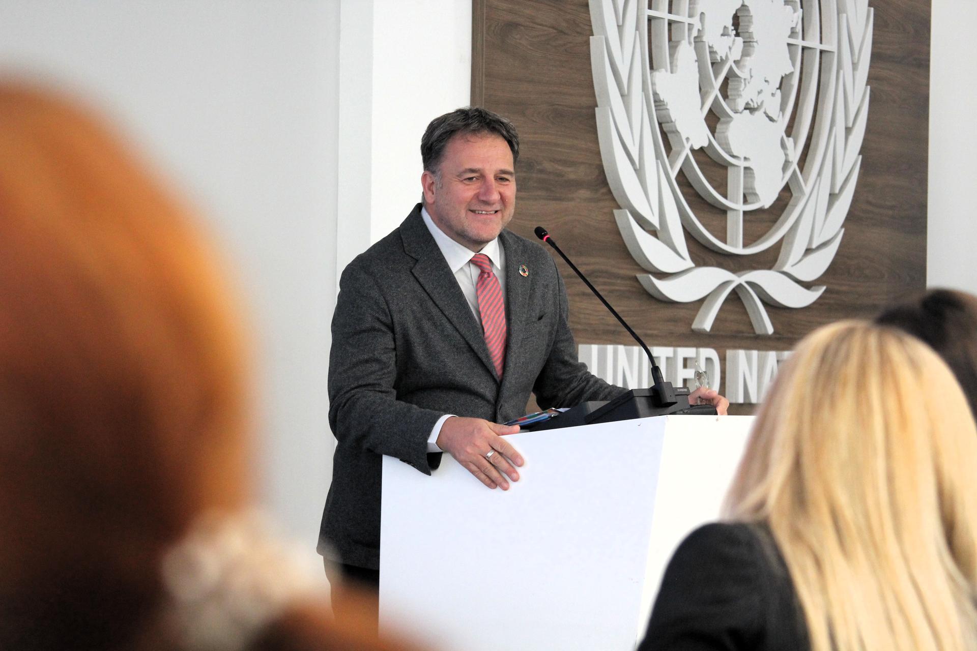 Photograph of a smiling man in a suit at a podium, UN emblem on wall, audience in foreground.