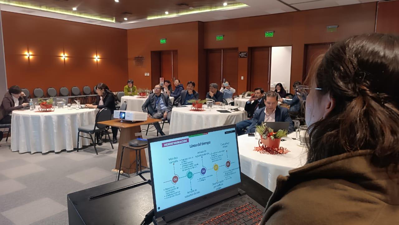 Photograph of a conference room; attendees at tables while a presenter speaks at the front.