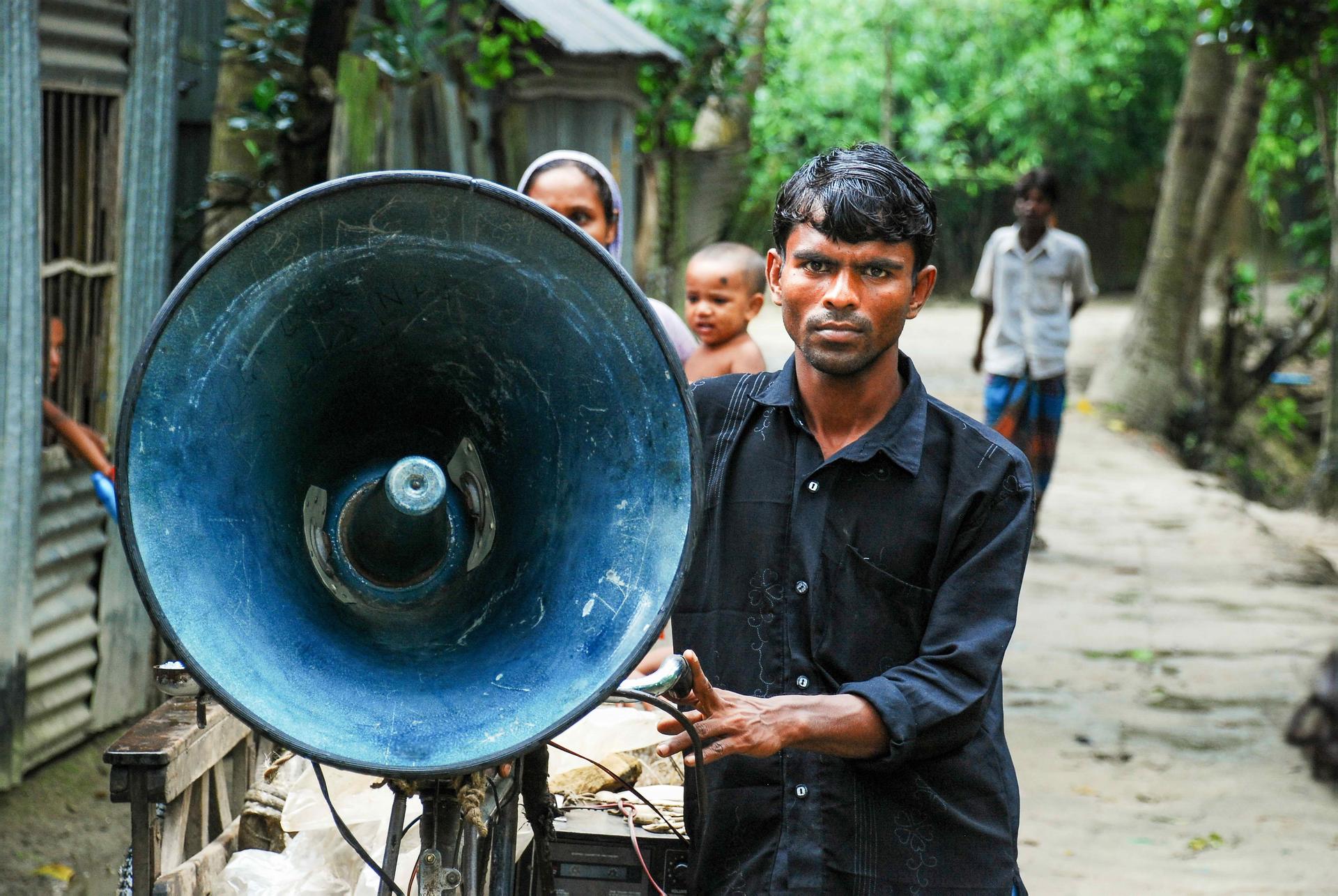 Man in a dark shirt uses a blue megaphone on a street; pedestrians in the background.