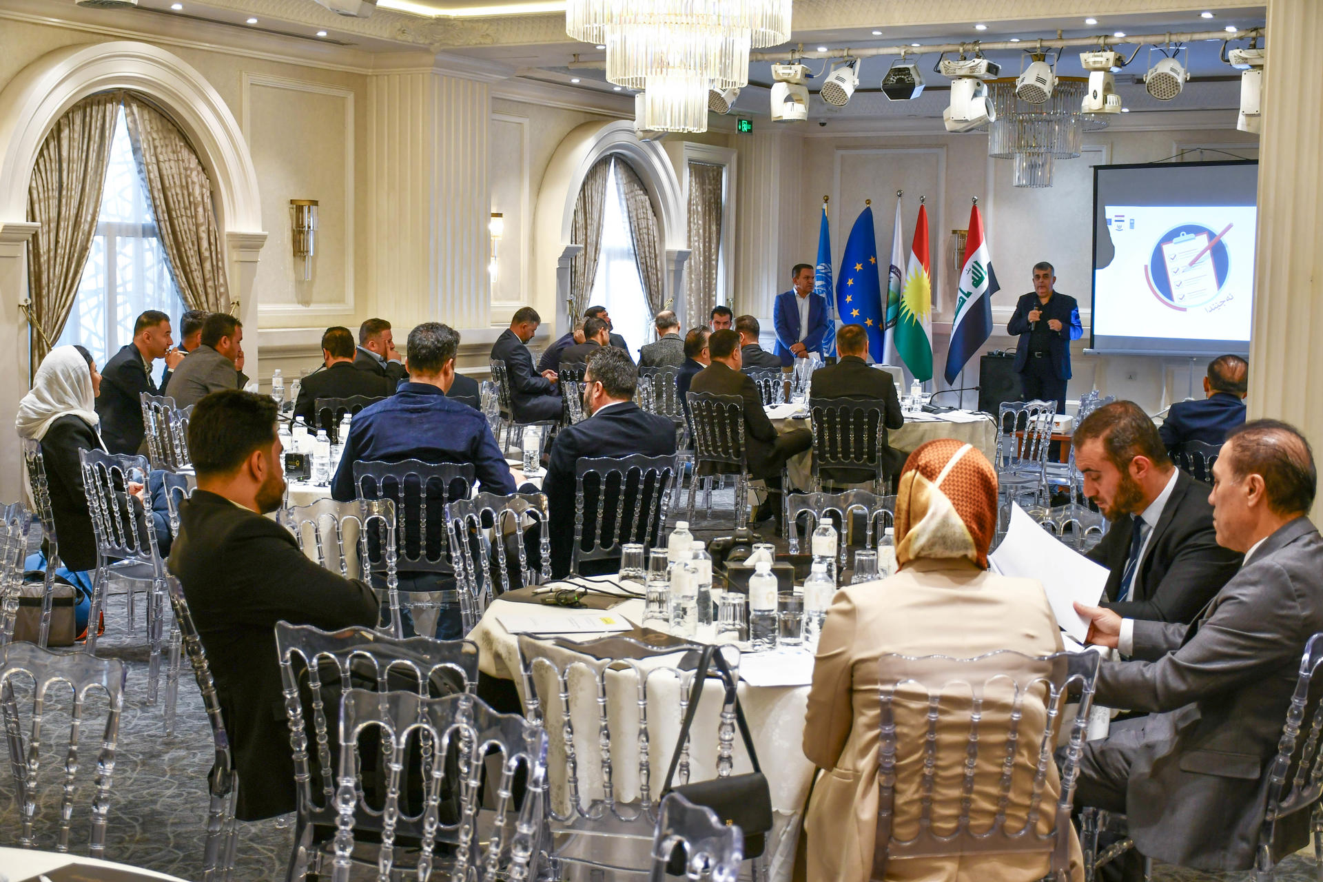 Conference room with attendees at round tables; presenter near screen with flags.