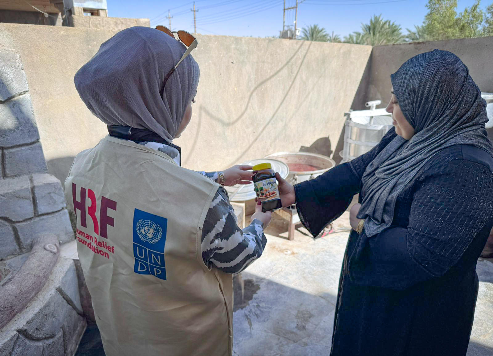 Two hijab-clad women exchange a bottle at a construction site; one wears a beige HRF jacket.