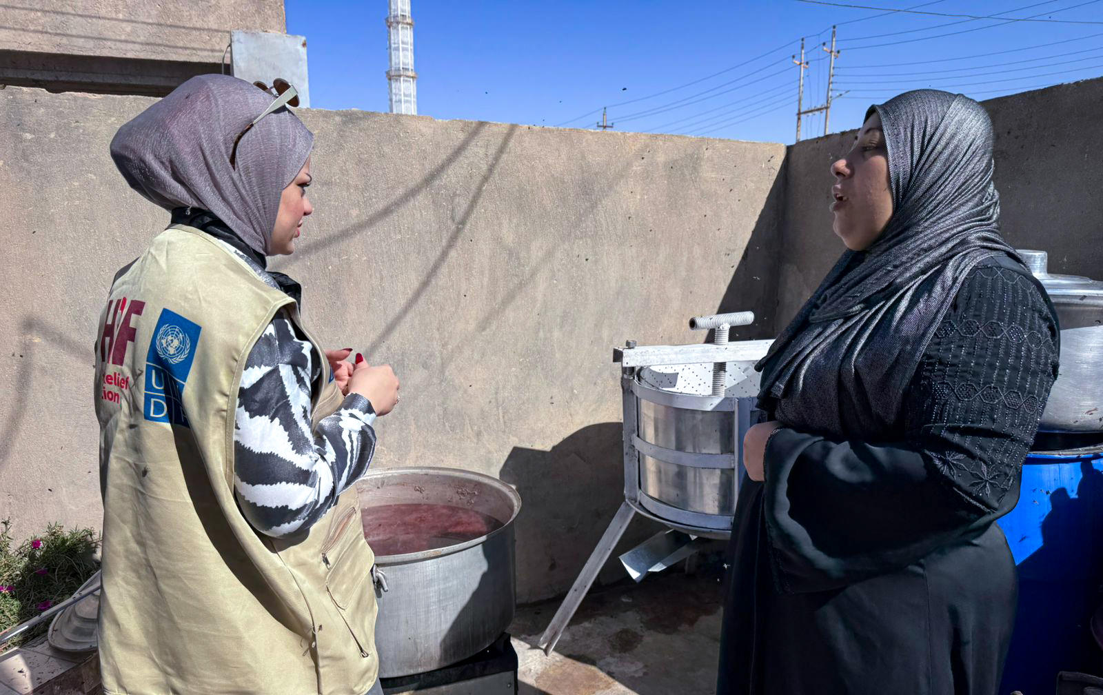 Photo: Two women wearing headscarves talk outdoors beside a blue water barrel.