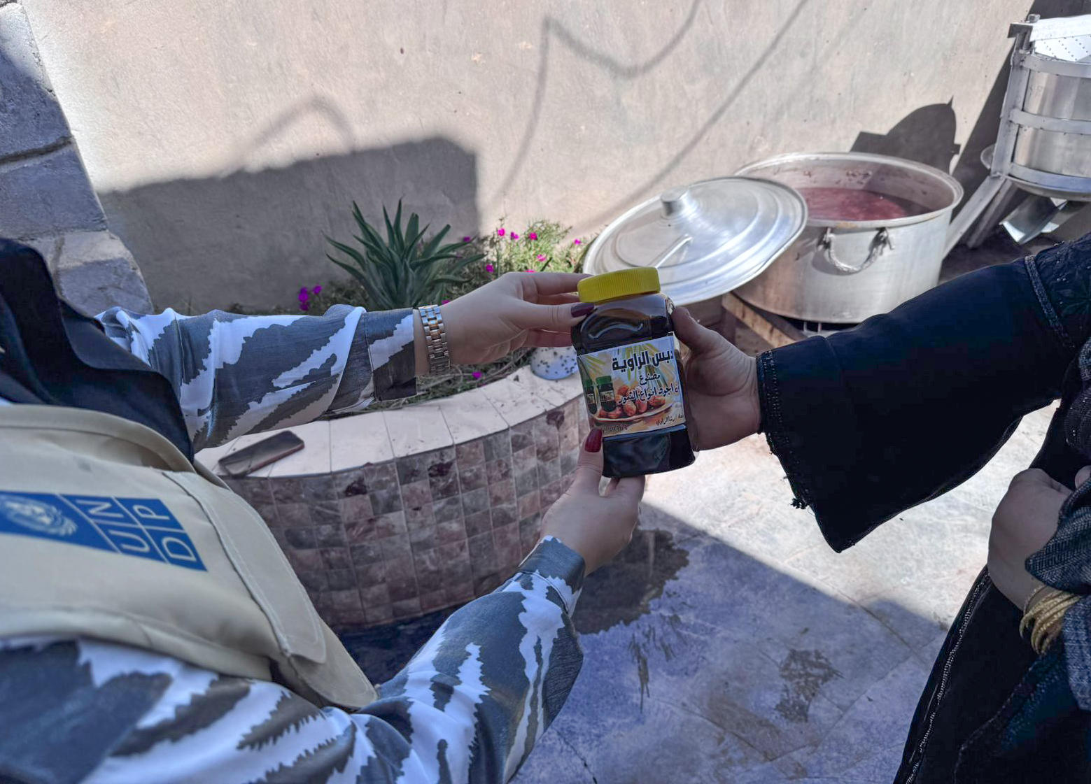 Two people exchange a colorful beverage container outdoors on a stone path.