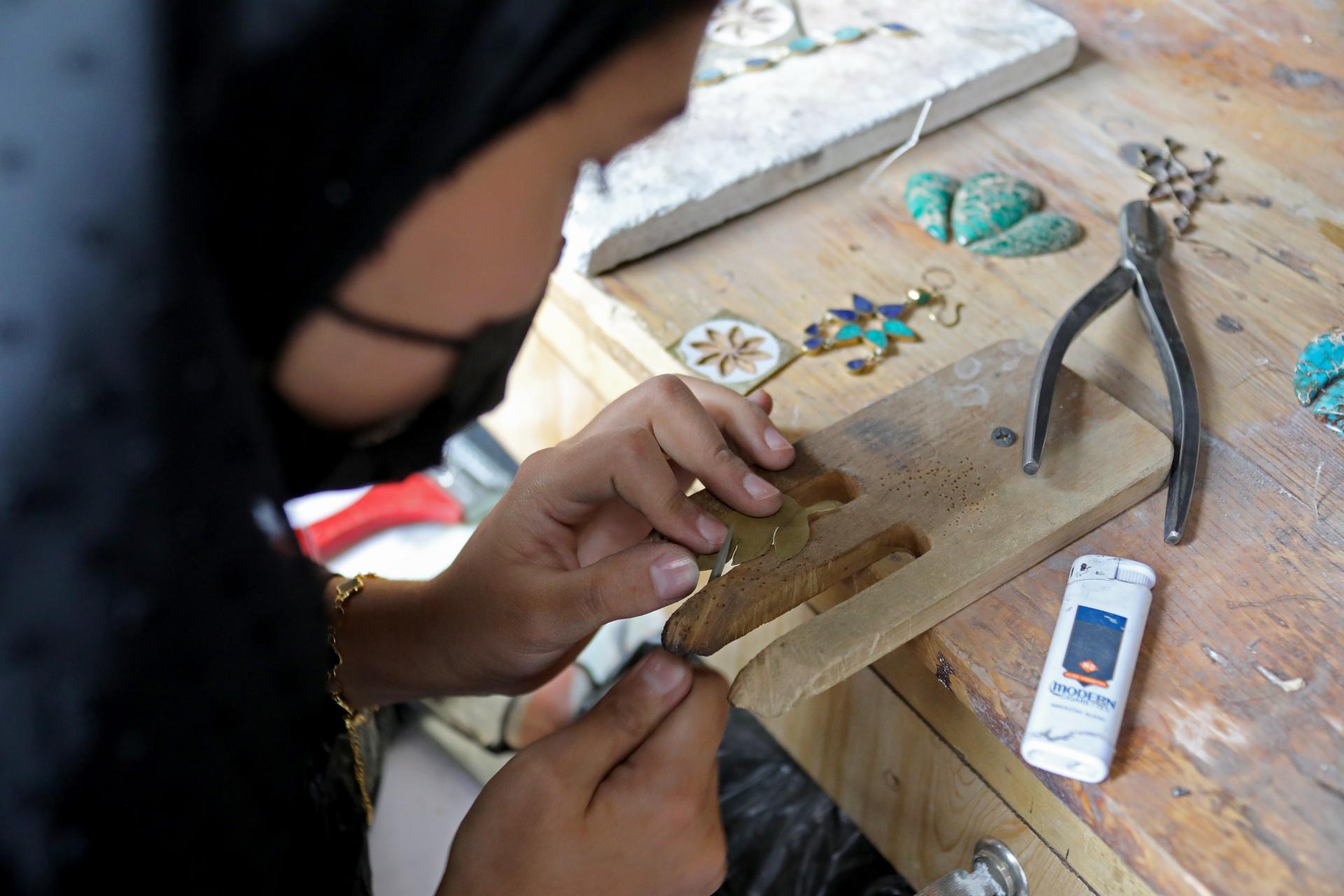 Hands crafting jewelry on a wooden workbench with beads, pliers, and a smartphone nearby.