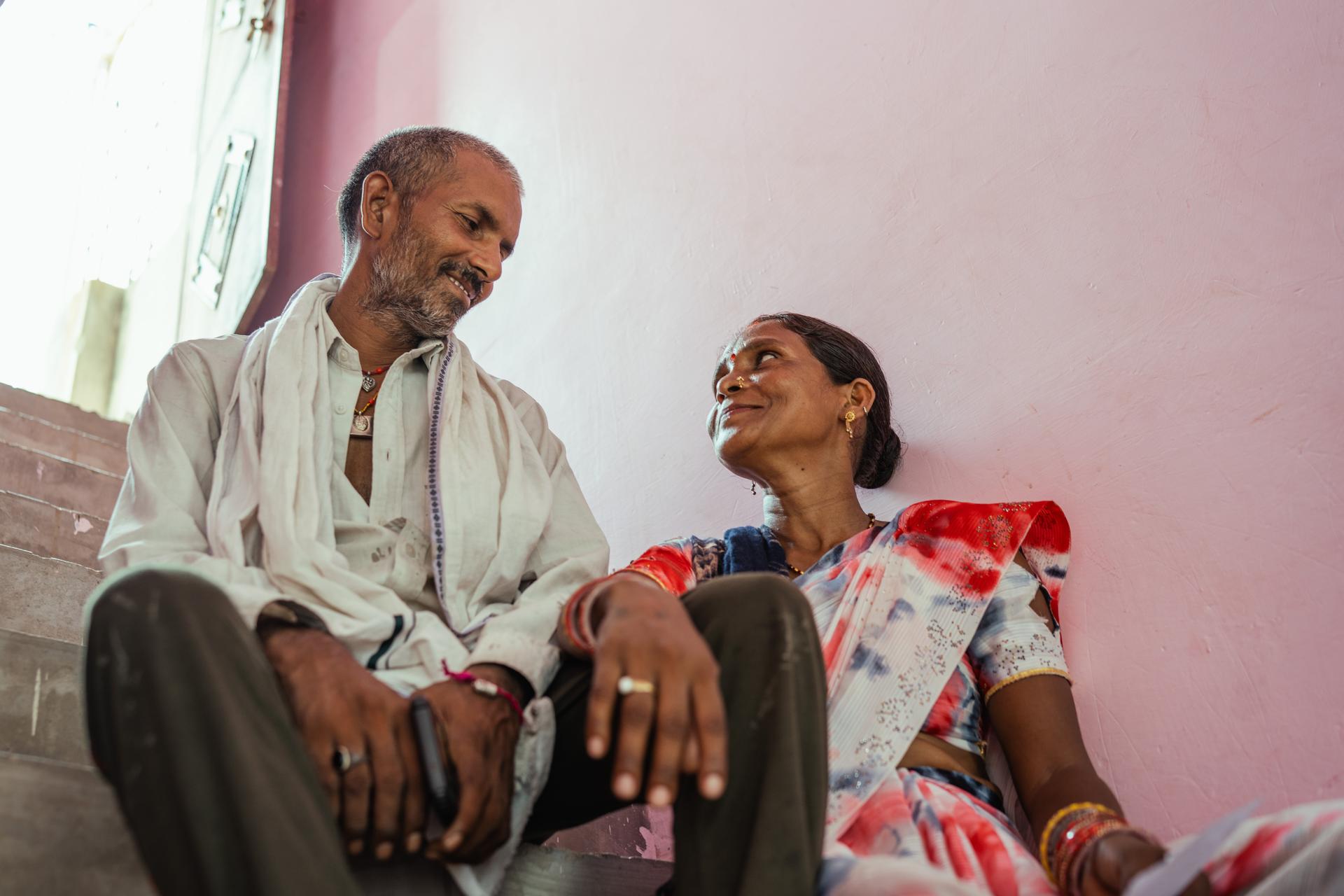 Two people sit and chat in a pink room; man in white shirt and tie, woman in a colorful dress.