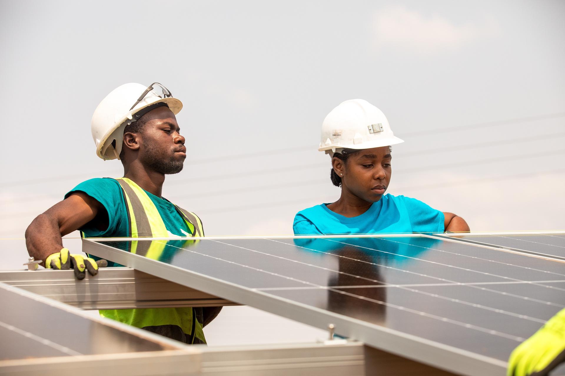 Two workers wearing hard hats inspect solar panels outdoors.
