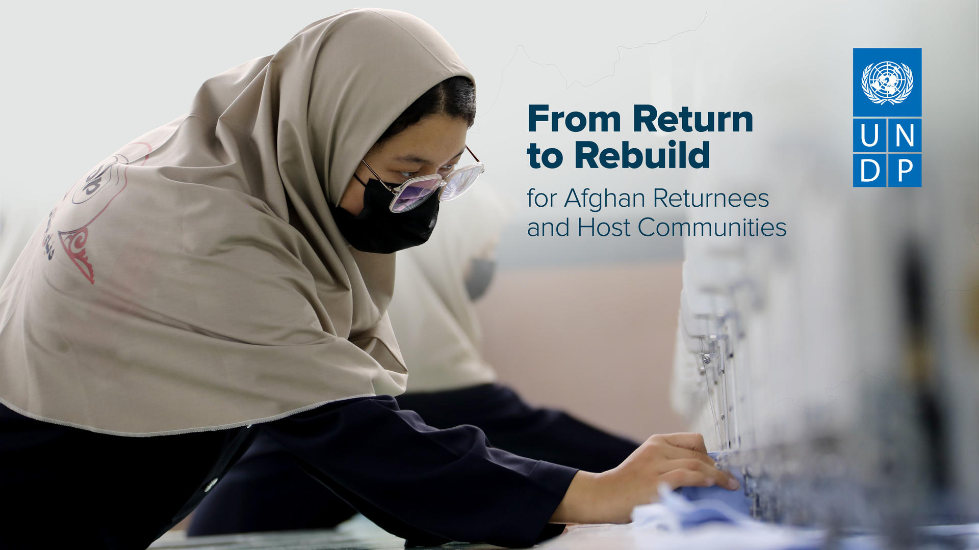 Photograph of a woman wearing a hijab and mask, writing at a desk; UNDP banner in background.