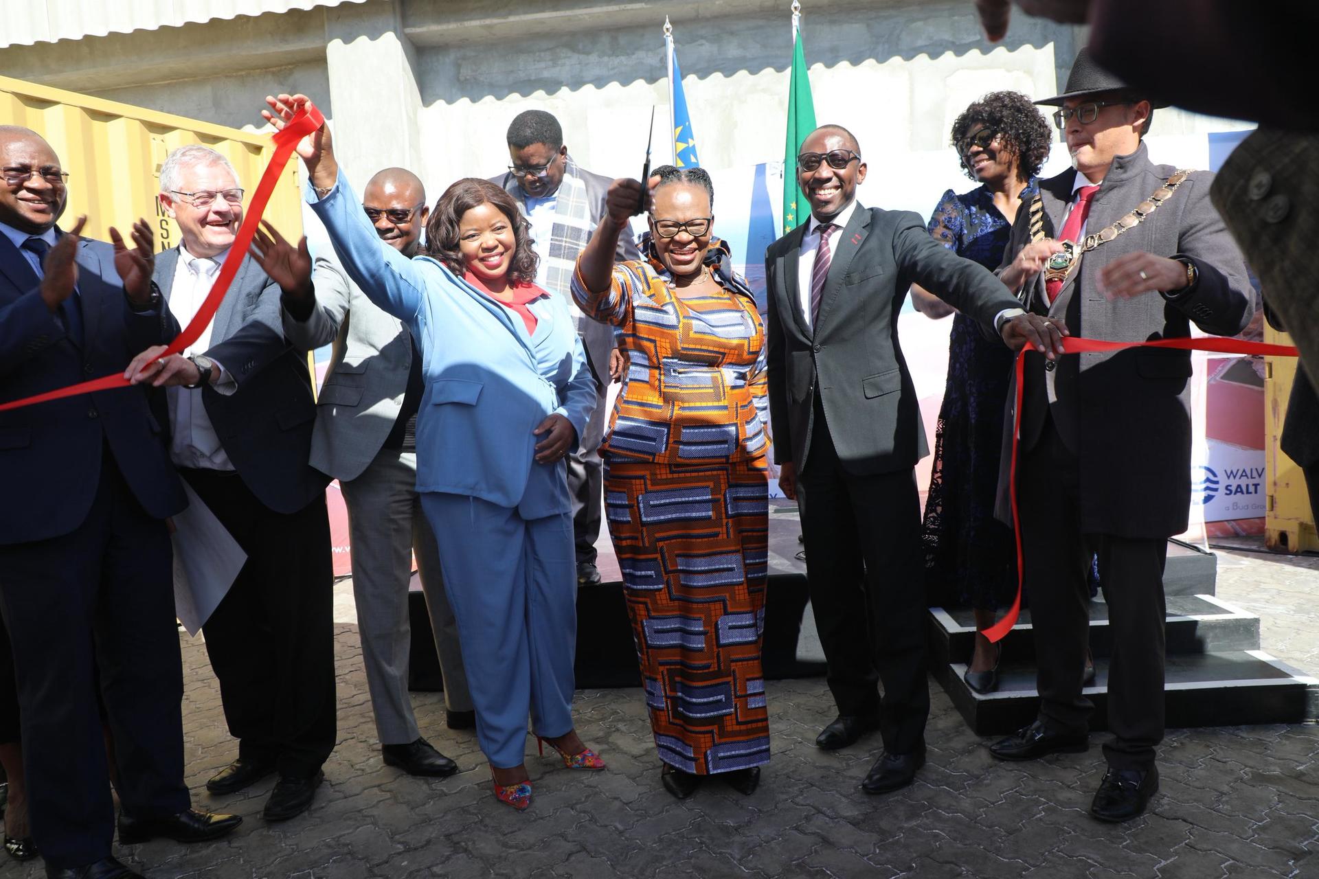 Diverse group of officials and community leaders cut a red ribbon at an outdoor ceremony.