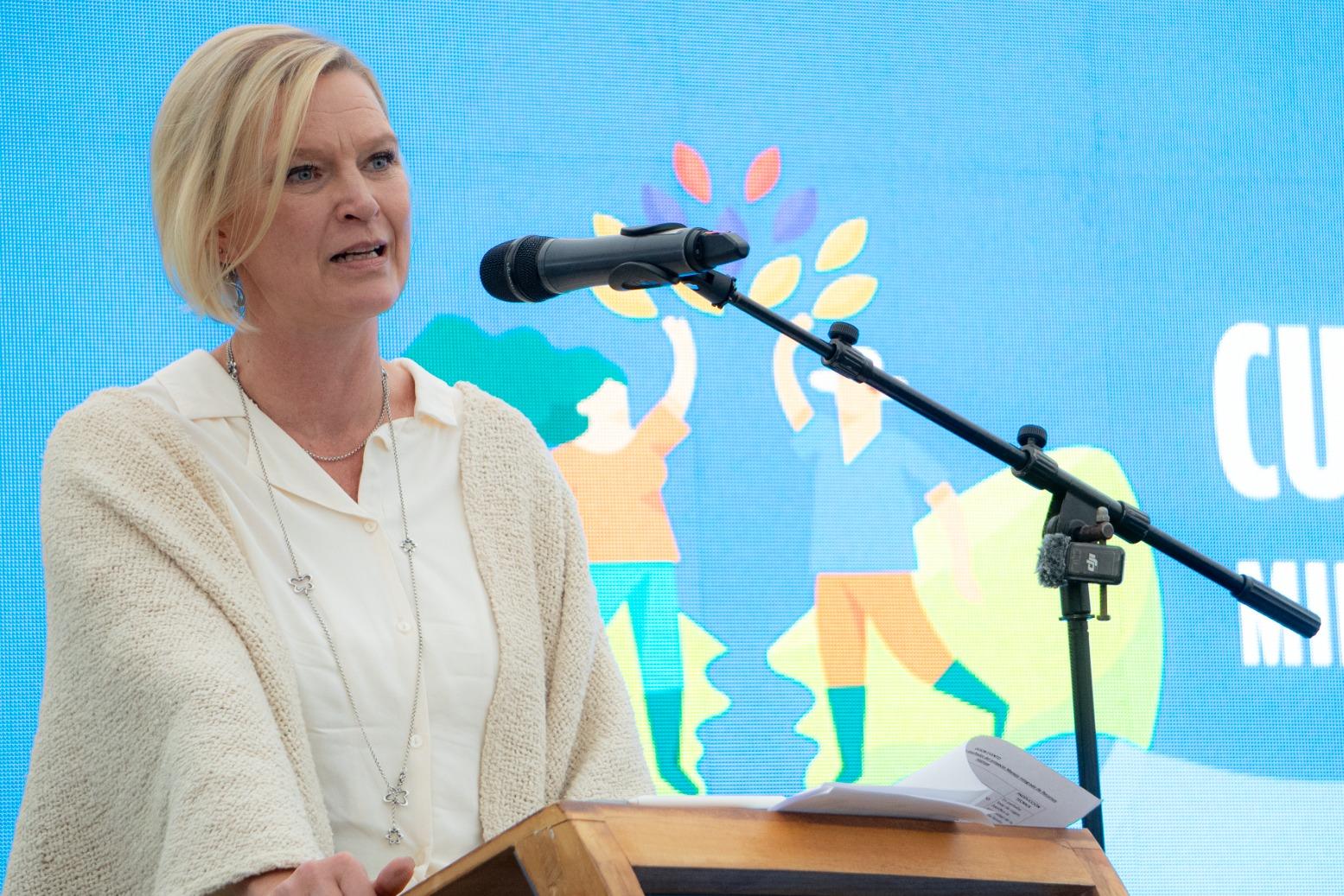Photo: Speaker at podium with microphone, wearing a light blazer, against a blue event backdrop.