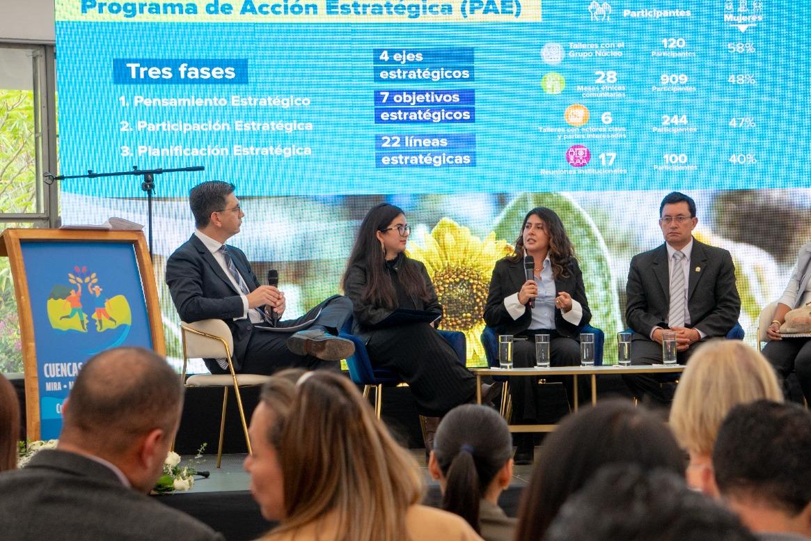 Panelists and moderator on stage at a conference, blue program screen behind, audience in front.
