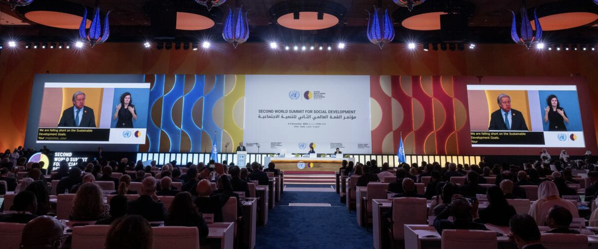 Large conference hall with a stage, central and side screens, colorful banners, and an audience.