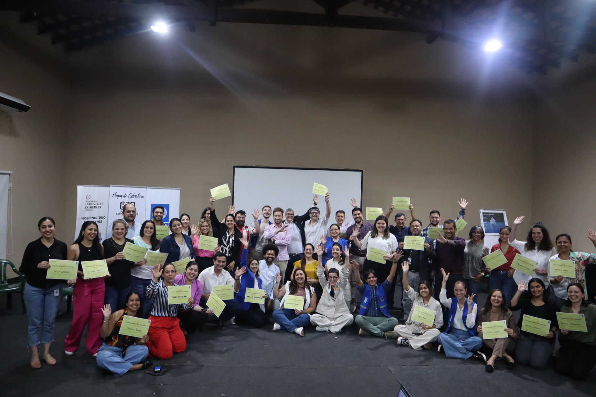 Group photo of diverse people on stage holding yellow papers, projector screen behind.