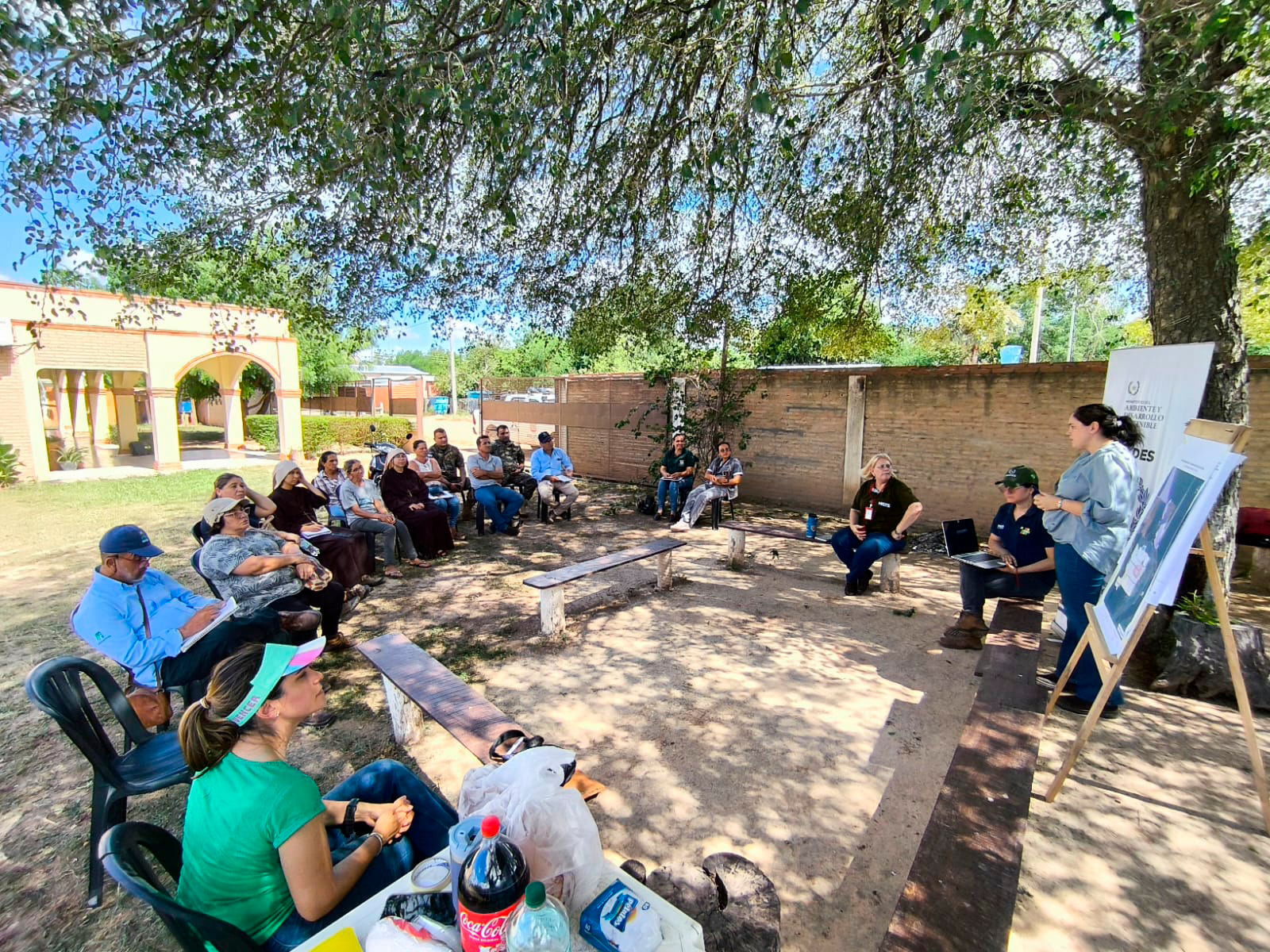 Outdoor group workshop with people seated in a circle under a tree; presenter with whiteboard.