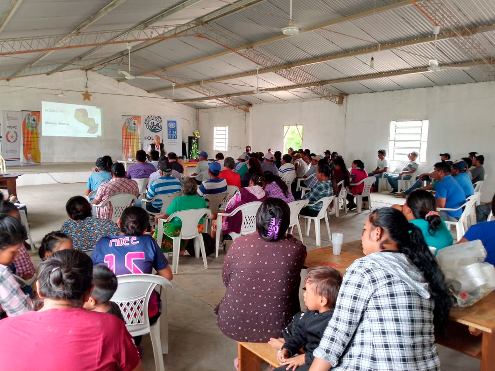 Crowded hall for a community meeting, attendees seated and watching a projector presentation.