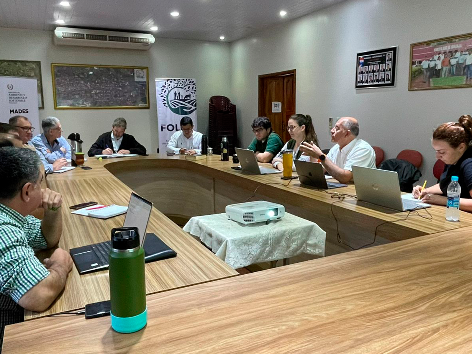 Group of people seated around a long wooden conference table in a bright meeting room.