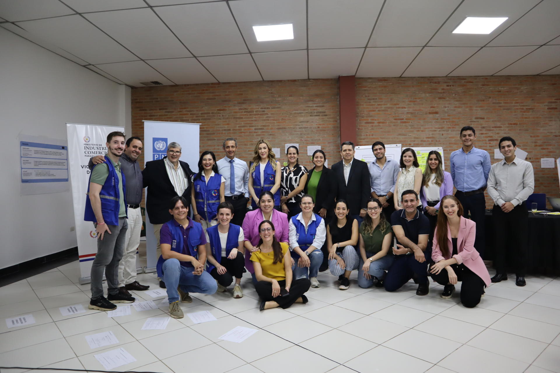 Group of diverse people posing for a group photo in a bright indoor lobby with a brick wall.
