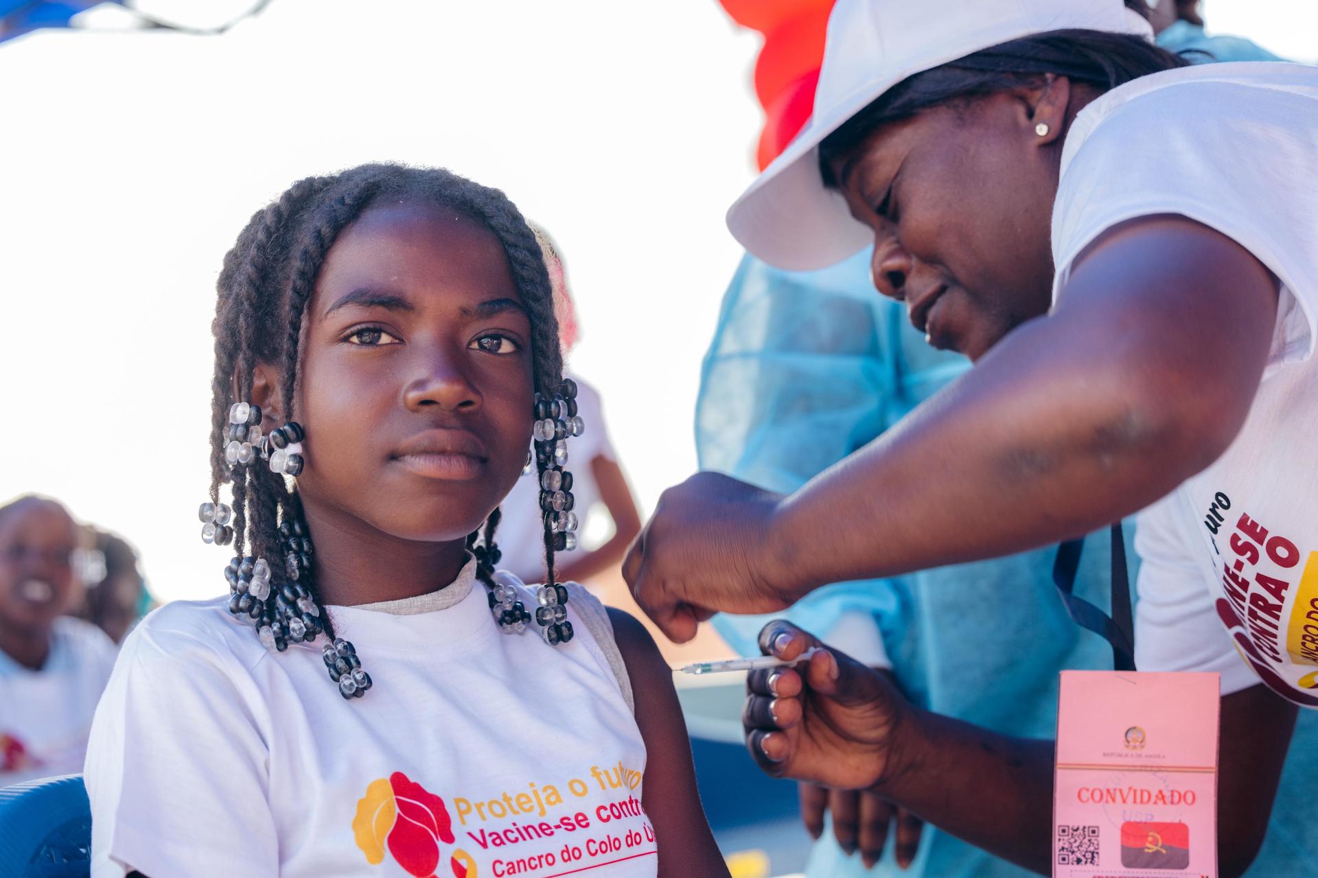 Girl with braided hair in a white charity shirt, next to a person with a pink badge outdoors.