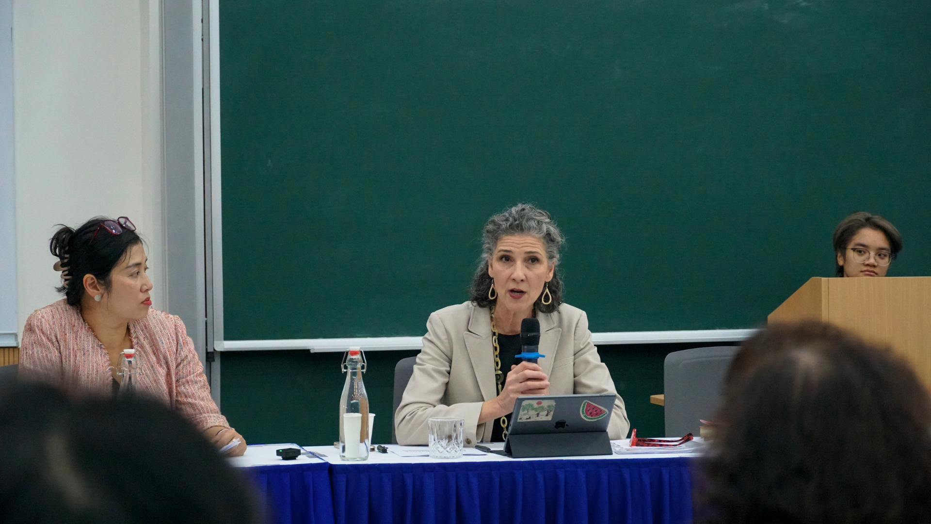 Panel discussion with three women at a table on stage, one speaking, green chalkboard behind.