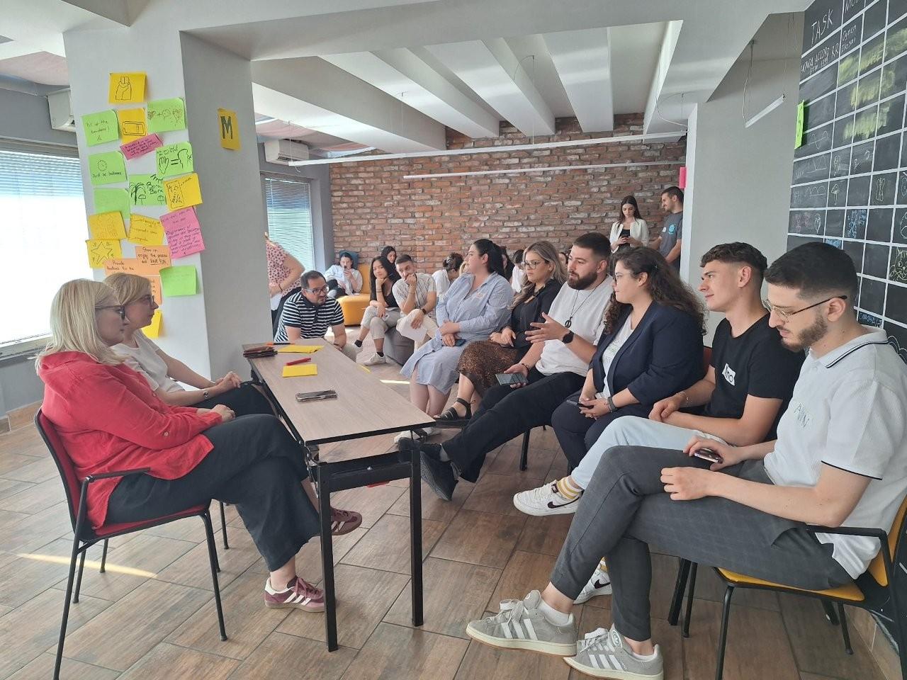Workshop scene: a woman in red leads discussion among seated participants with colorful notes.