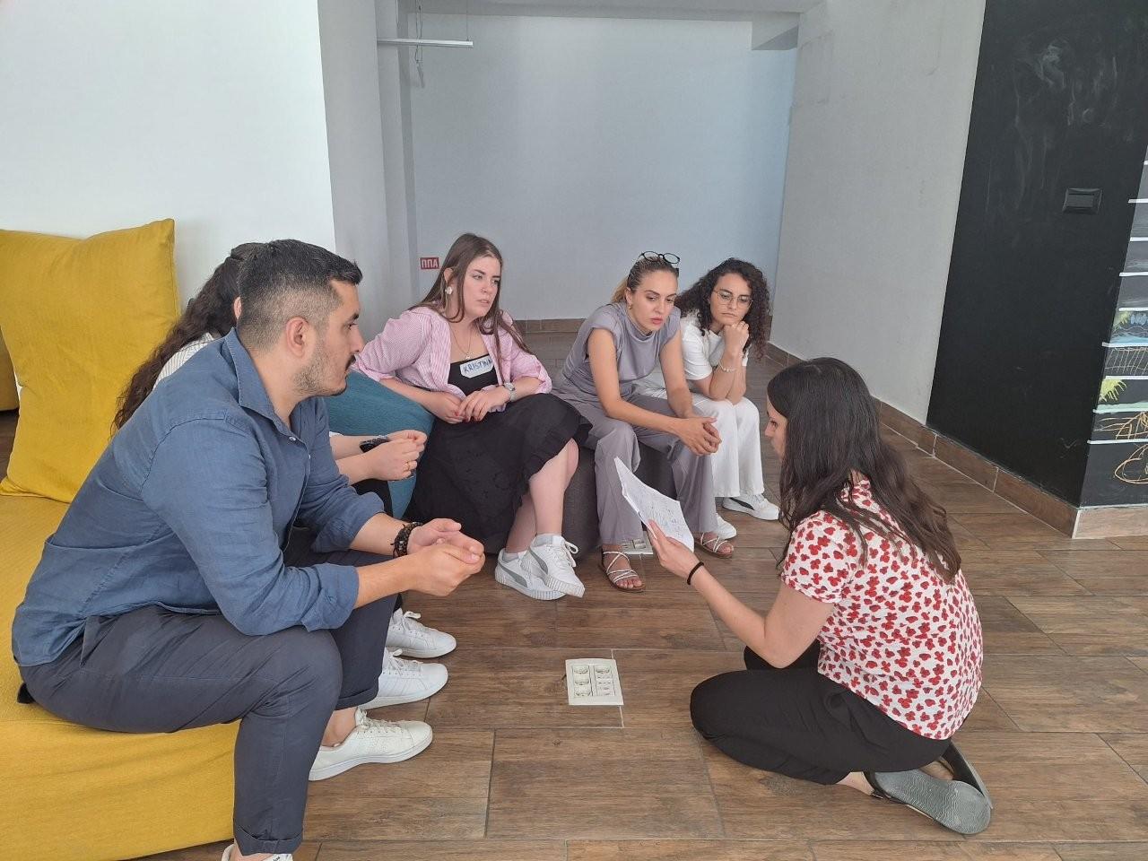 Six people sit on cushions around a wooden floor, chatting in a living room.